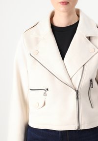 Cropped white biker jacket in soft fabric, featuring two zippered pockets, asymmetrical zipper closure, and snap collar details.