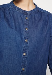 Primo piano di una persona che indossa una camicia di jeans blu con colletto rotondo e cinque bottoni visibili sulla parte frontale.