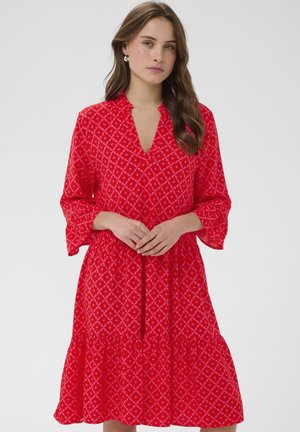 Žena s dlhými hnedými vlasmi, oblečená v červenom šate s vzorom, s trojštvrťovými rukávmi a výstrihom do V, stojaca so zloženými rukami.