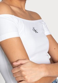 Žena nosící bílý off-shoulder top Calvin Klein Jeans, s crossed arms, na jednoduchém světlém pozadí.