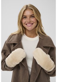 Smiling woman with blonde hair wears beige faux fur mittens, brown wool coat, and white sweater against plain light gray background.