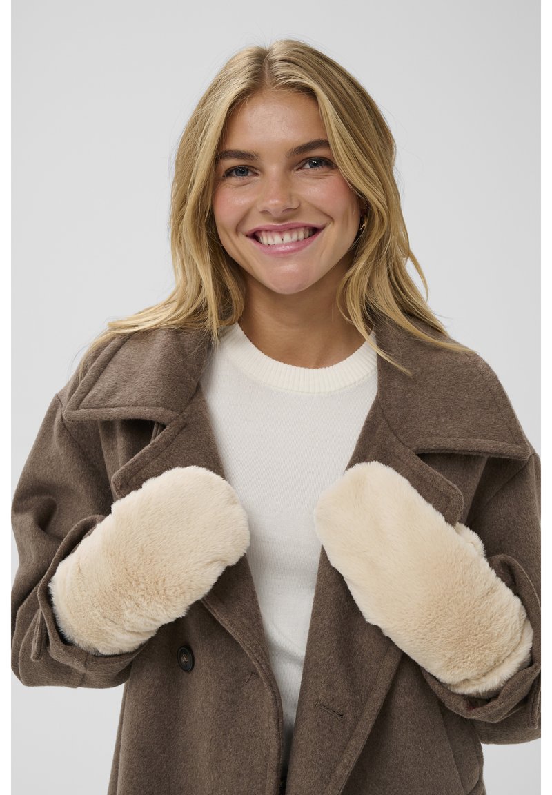 Smiling woman with blonde hair wears beige faux fur mittens, brown wool coat, and white sweater against plain light gray background.