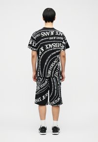 Άτομο με κοντά μαύρα μαλλιά που φοράει μαύρο σορτς και μπλούζα με λευκό μοτίβο από στροβιλιζόμενο κείμενο "Versace Jeans Couture", στέκεται μπροστά σε λευκό φόντο.