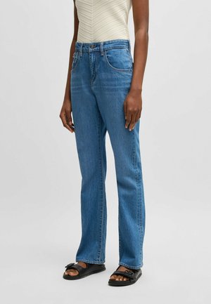 Blaue Denim-Jeans mit geradem Bein, featuring einen hohen Bund, Fünf-Taschen-Design und dezente Ausbleichungen. Tragen mit schwarzen Riemchensandalen.