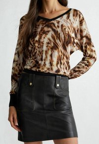Langärmeliges V-Ausschnitt-Oberteil mit braun-cremigem abstraktem Muster, kombiniert mit einem schwarzen Leder-Minirock mit Fronttaschen und Druckknöpfen.