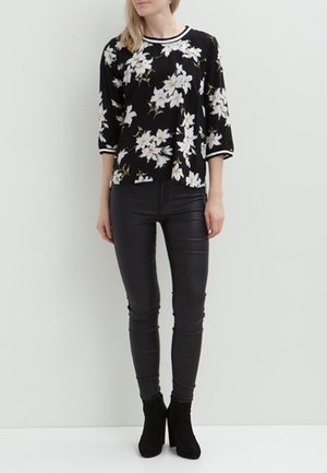 Kobieta ubrana w czarną kwiecistą bluzkę z rękawami o długości 3/4, czarne dopasowane spodnie i czarne botki na wysokim obcasie, stojąca na tle białego, jednolitego tła.