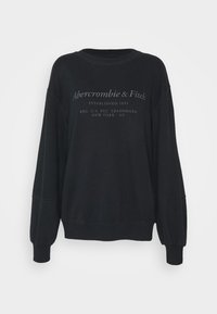 Sudadera negra con cuello redondo. Presenta mangas largas y ligeramente abullonadas. Muestra texto blanco, un logotipo y la fecha de establecimiento en la parte frontal.