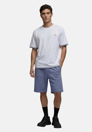 Homme portant une chemise blanche à manches courtes avec une bordure rayée et un short bleu, debout avec une main dans la poche devant un fond uni.