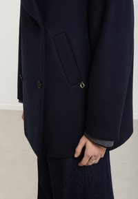 Námornícky dvojradový kabát z textúrovanej látky, s bočnou vreckom a elegantným límcom, skombinovaný so šedým svetrom pod ním.