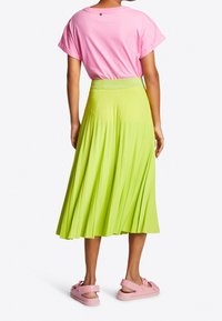 Kvinna som står vänd bort och bär en rosa t-shirt, limegrön plisserad midi kjol och rosa sandaler med öppen bakdel och rosetter.