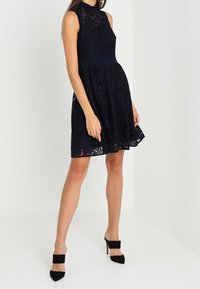 Ärmlös marinblå spetsklänning med hög halsringning och figurnära midja, matchad med svarta spetsiga klackskor med breda remmar.