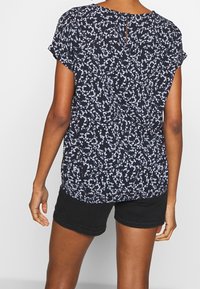 Blusa blu navy con stampa floreale bianca, maniche corte, scollatura rotonda e dettaglio a chiave sul retro, abbinata a pantaloni corti neri.