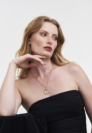 Gouden hangerketting met een letterdetail, gedragen op een blote schouder, gecombineerd met een nauwsluitende zwarte strapless top. Glad oppervlak en elegant ontwerp.