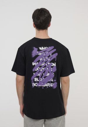 T-shirt nera in cotone con design di vernice spray viola sul retro con testo bianco. Maniche corte, vestibilità comoda, scollatura rotonda semplice.