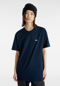 Un jeune portant un bonnet en tricot noir, des colliers en argent superposés, et un t-shirt Vans oversized bleu marine avec un pantalon sombre sur fond blanc.