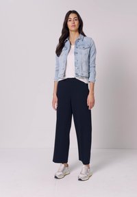 Donna in piedi che indossa una giacca di jeans azzurro chiaro, una camicia bianca, pantaloni neri a gamba larga e sneakers bianche su uno sfondo uniforme.