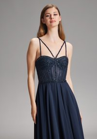 Abito da sera blu navy con corpetto decorato di perline, scollatura trasparente e spalline sottili. La gonna scorre elegantemente dalla vita, realizzata in un tessuto leggero.
