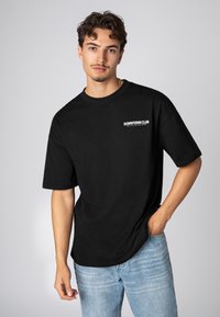 Schwarzes Baumwoll-T-Shirt mit lockerer Passform, rundem Halsausschnitt und kurzen Ärmeln. Mit weißem "DOWNTOWN CLUB"-Text auf der linken Brustseite.
