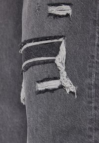 Detailní záběr šedé džínové látky s několika obdélníkovými opotřebovanými a třepenými záplatami odhalujícími bílé nitě pod látkou.