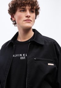 Νεαρό άτομο με σγουρά μαλλιά που φορά μαύρο μπουφάν Calvin Klein και μαύρο T-shirt με λευκό κείμενο σε απλό φόντο.