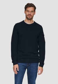 Marineblauwe crewneck sweatshirt met ribbelmanchetten en -zoom; gestructureerde stof; heeft een mouwzak met een donkere patch. G gedragen over spijkerbroeken.