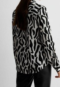 Donna che indossa una blusa a maniche lunghe con un motivo astratto bianco e nero abbinata a pantaloni neri, vista da dietro su uno sfondo neutro.