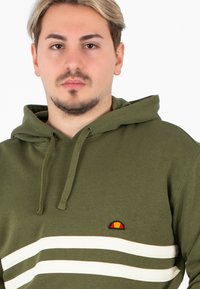 Felpa verde oliva con tasca frontale, caratterizzata da strisce bianche orizzontali e un piccolo logo ricamato in rosso e giallo. Cappuccio con cordini.