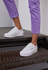 Baskets blanches à lacets avec une semelle en caoutchouc texturé, un accent argenté au talon et un logo de marque sur le côté. Portées avec un jean lavande.