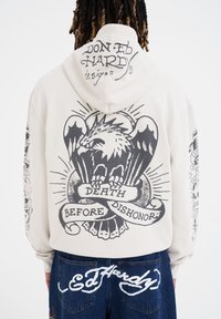 Sudadera de color crema con un gran diseño gráfico en negro de un pájaro y un pergamino con el texto "MUERTE ANTES QUE DESHONRA" en la parte posterior.