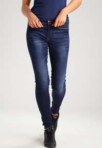 Sötétkék farmer skinny farmernadrág, finom kopásokkal, öt zsebes kialakítással és kontrasztos varrással. Fekete sportcipővel párosítva.