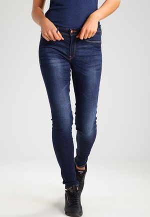 Schmale Jeans aus dunkelblauem Denim mit sanften Verblassungen, fünf Taschen und kontrastierender Naht. Kombiniert mit schwarzen Sportschuhen.