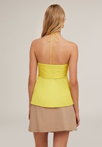 Geel geplooide haltertop met een aansluitende buste, verstelbare strik om de hals en een uitlopende zoom, gecombineerd met een beige rok. Soepele textuur van de stof.