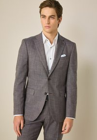 Smeđi kaput u houndstooth uzorku s dva dugmeta, izrezom sa strane i bočnim džepovima. Bijela košulja i maramica u džepu dopunjuju odjevnu kombinaciju.