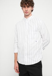 Balta pogājama krekla ar zilām vertikālām svītrām, garām piedurknēm, vienu krūšu kabatu un smalku logotipa detaļu. Kokvilnas auduma tekstūra.