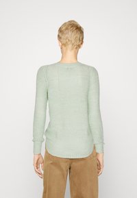 Osoba s krátkými blond vlasy, oblečená v světle zeleném pleteném svetru a hnědých kalhotách, viděná zezadu na neutrálním pozadí.