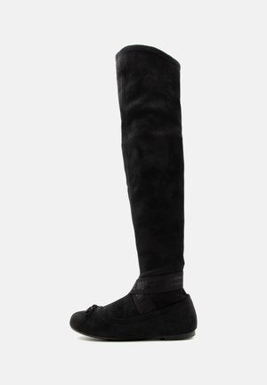 Schwarzer flacher Overknee-Stiefel mit abgerundetem Zehenbereich, weichem Material und dekorativem Riemen um den Knöchel vor einem schlichten hellen Hintergrund.