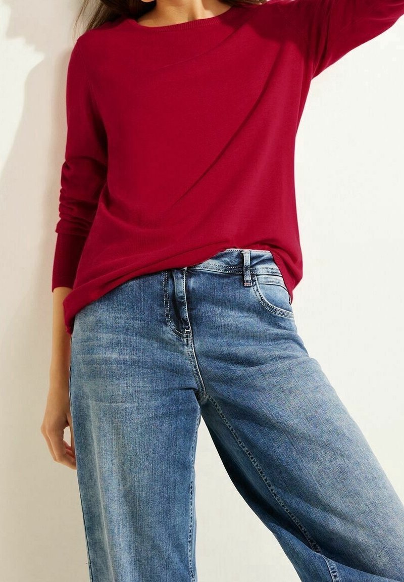 En person som bär en röd långärmad tröja som är lätt instoppad i högmidjade, löst sittande blå jeans mot en ljus bakgrund.