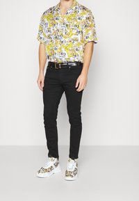 Mies, joka käyttää lyhythihaista valkoista paitaa, jossa on keltainen barokkikuvio, mustia slim-fit-housuja, mustaa vyötä ja valkoisia lenkkareita, joissa on samanlainen kuosi.