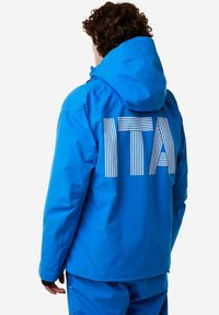 Persona con capelli ricci che indossa una giacca e pantaloni blu, in piedi con le spalle rivolte verso la camera; testo bianco a righe "ITA" sulla parte posteriore della giacca.