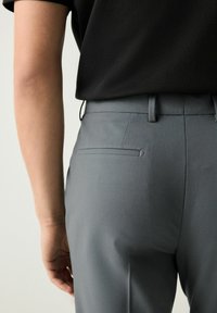 Pantalones grises ajustados con una textura suave y un bolsillo trasero. Llevados con una blusa negra, vistos de lado y por detrás.