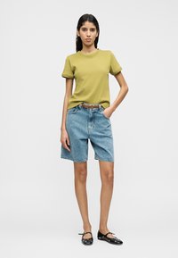 Πράσινο κοντομάνικο t-shirt με στρογγυλή λαιμόκοψη, συνδυασμένο με ανοιχτό μπλε τζιν σορτς. Το μοντέλο φοράει μαύρα μπαλαρίνες και καφέ ζώνη.