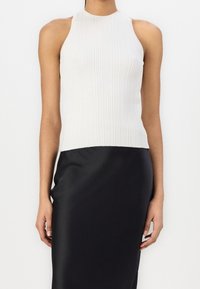 Mujer que lleva un top de halter blanco de canalé sin mangas y una falda negra de cintura alta y suave, de pie contra un fondo claro y liso.