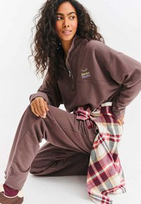Brązowa bluza polarowa na zamek z haftem "Naoko Studio", w komplecie z dopasowanymi spodniami. Koszula w kratę w kolorach czerwonym, białym i niebieskim zawiązana w talii.
