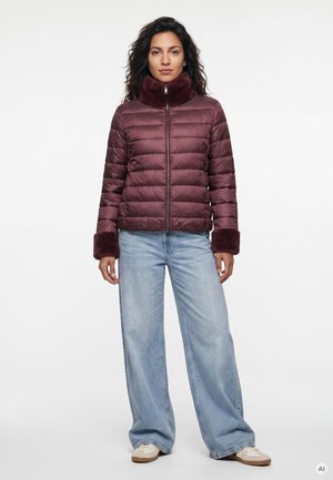 Frau mit dunklem Haar, die eine burgunderrote Steppjacke mit Fellmanschetten und Kragen, helle weite Jeans und weiße Sneakers trägt, steht vor weißem Hintergrund.
