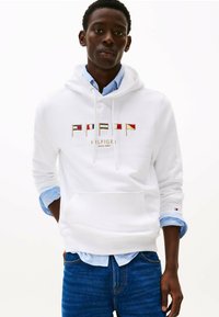 Witte capuchontrui met geborduurd logo dat rode, blauwe en gouden vlaggen op de borst heeft. De stof lijkt zacht; vooraan een buidelzak.