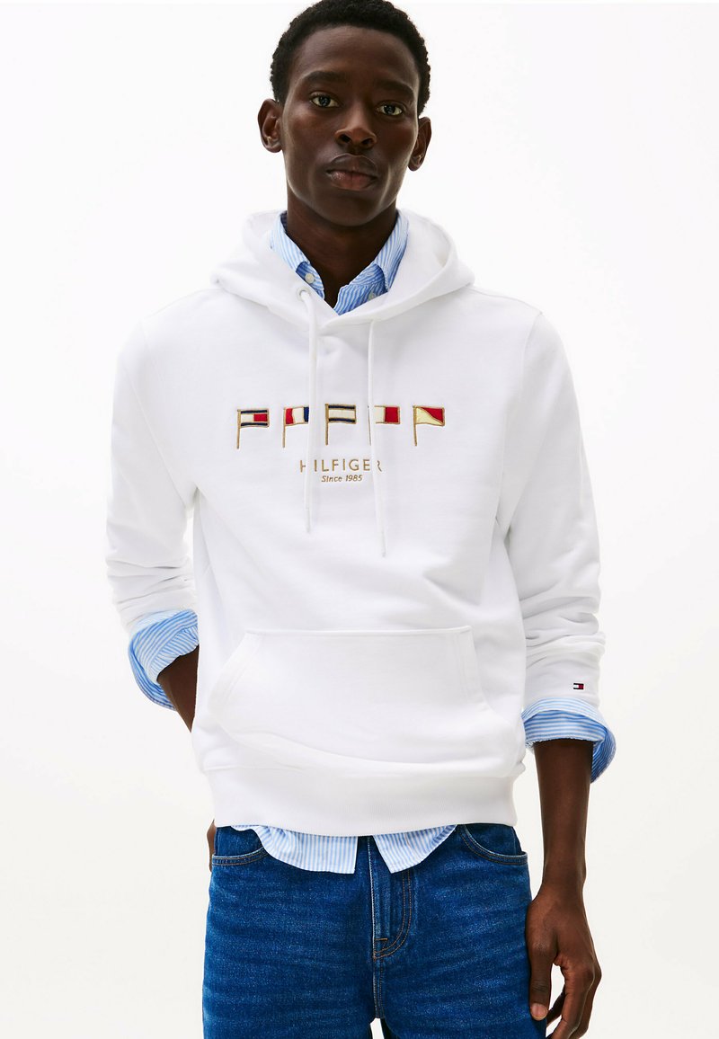 Witte capuchontrui met geborduurd logo dat rode, blauwe en gouden vlaggen op de borst heeft. De stof lijkt zacht; vooraan een buidelzak.
