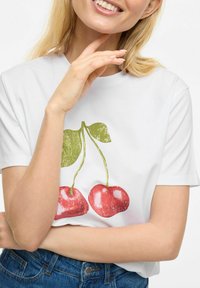 T-shirt blanc avec un motif de deux cerises rouges accompagnées de feuilles vertes. Matière en coton, manches courtes, coupe décontractée, associé à un jean bleu.