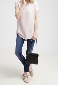 Světle růžová blouse s krátkým rukávem a zakřiveným lemem, v kombinaci s modrými džíny a béžovými botami. Drží černou ramenní tašku s jednoduchým designem.