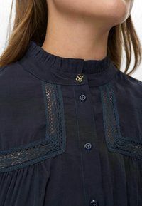 Woman wearing dark blue blouse with pleated high collar, lace trim details, front buttons, and a small gold flower-shaped pin near the collar.