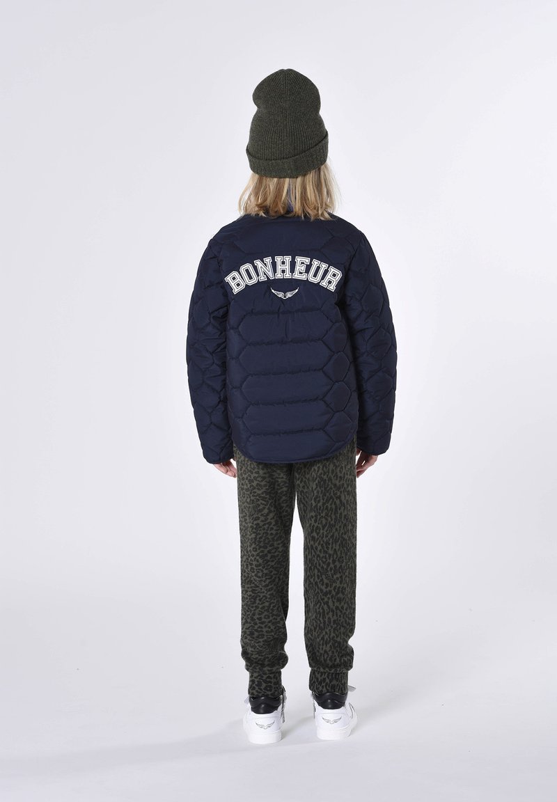 Veste matelassée marine avec le texte "BONHEUR" au dos, pantalon vert léopard, et un bonnet en tricot vert, associé à des baskets blanches.